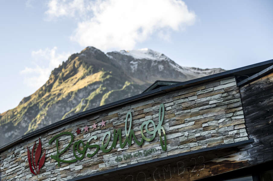 Hoteleingang mit Logo, im Hintergrund ein Berggipfel, blauer Himmel und 1 Wolke